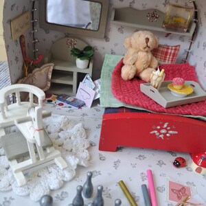 Op de afbeelding: Een gedetailleerde miniatuur poppenhuis sc&egrave;ne met een rood bed, een teddybeer en een dienblad met een cupcake en een ijsje. De sc&egrave;ne bevat een schommelpaard, kegels en potloden, allemaal op een bloemmotief oppervlak.