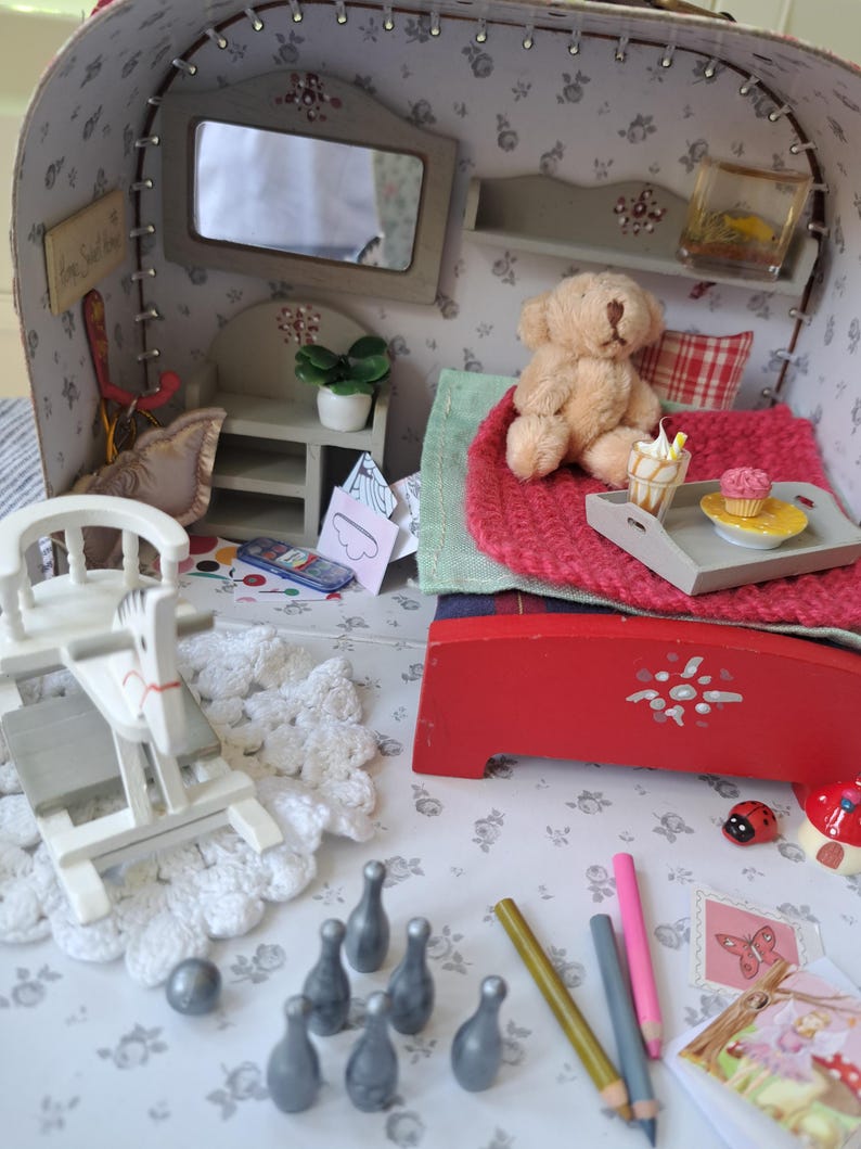 Op de afbeelding: Een gedetailleerde miniatuur poppenhuis sc&egrave;ne met een rood bed, een teddybeer en een dienblad met een cupcake en een ijsje. De sc&egrave;ne bevat een schommelpaard, kegels en potloden, allemaal op een bloemmotief oppervlak.