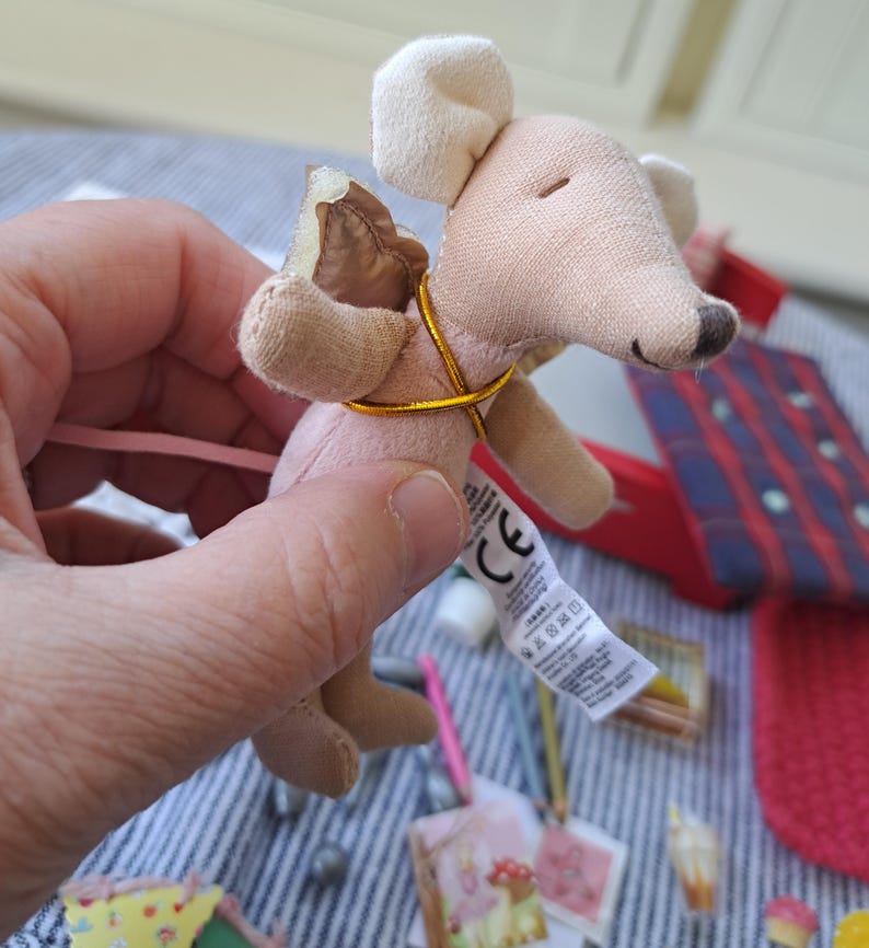 Op de afbeelding: Een kleine, pluche muis speelgoed in roze en beige, met een goudkleurig koord om de nek. De muis heeft een lange, roze staart en een label bevestigd. Het speelgoed wordt in een hand gehouden, met andere kleine items op de achtergrond.