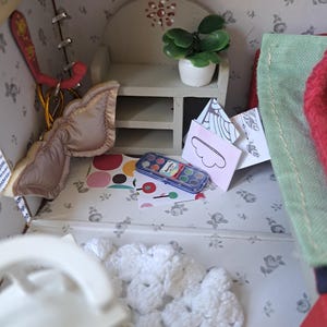 Op de afbeelding: Een gedetailleerd miniatuur poppenhuisinterieur met een wit gehaakt kleed, een kleine plant in een pot en een aquarelverfset. De muren zijn versierd met een bloemenpatroon. Een klein bordje met de tekst "Sweet home" is zichtbaar.