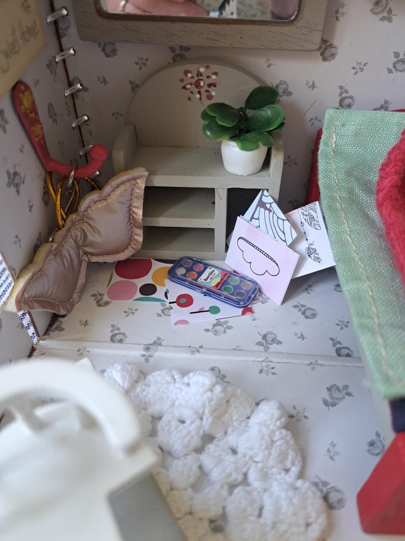 Op de afbeelding: Een gedetailleerd miniatuur poppenhuisinterieur met een wit gehaakt kleed, een kleine plant in een pot en een aquarelverfset. De muren zijn versierd met een bloemenpatroon. Een klein bordje met de tekst "Sweet home" is zichtbaar.