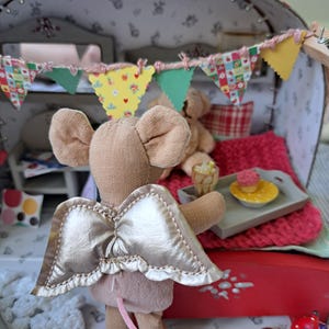 Op de afbeelding: Een miniatuur poppenhuis sc&egrave;ne met een beige muis met gouden vleugels. De muis staat voor een rood bed met een roze deken. Een klein dienblad bevat een cupcake en een drankje. Een kleurrijke banner hangt erboven.