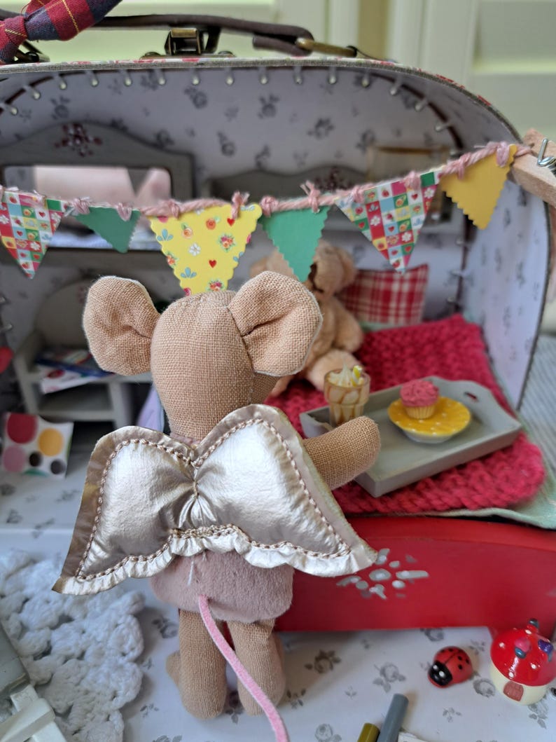 Op de afbeelding: Een miniatuur poppenhuis sc&egrave;ne met een beige muis met gouden vleugels. De muis staat voor een rood bed met een roze deken. Een klein dienblad bevat een cupcake en een drankje. Een kleurrijke banner hangt erboven.
