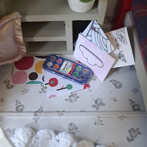 Op de afbeelding: Een miniatuursc&egrave;ne met een aquarelverfset, kleurrijke papieren en decoratieve kaarten. Een kleine witte kast met een plant in een pot staat op de achtergrond. De sc&egrave;ne is geplaatst op een bloemmotief.