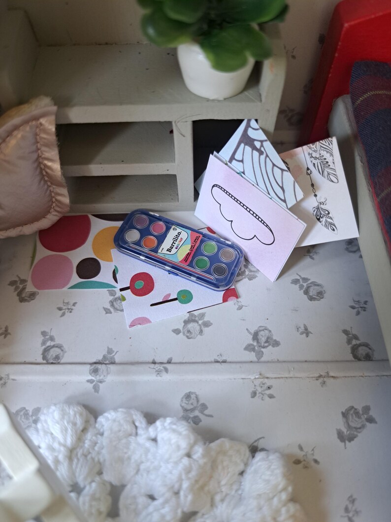Op de afbeelding: Een miniatuursc&egrave;ne met een aquarelverfset, kleurrijke papieren en decoratieve kaarten. Een kleine witte kast met een plant in een pot staat op de achtergrond. De sc&egrave;ne is geplaatst op een bloemmotief.