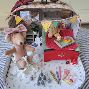 Op de afbeelding: Een miniatuur poppenhuis koffer met een bloemenpatroon, open om een gezellig interieur te onthullen. Een muispop met een roze strik zit in een miniatuurstoel. Het interieur omvat een bed, een teddybeer en miniatuuraccessoires zoals een dienblad met een cupcake.
