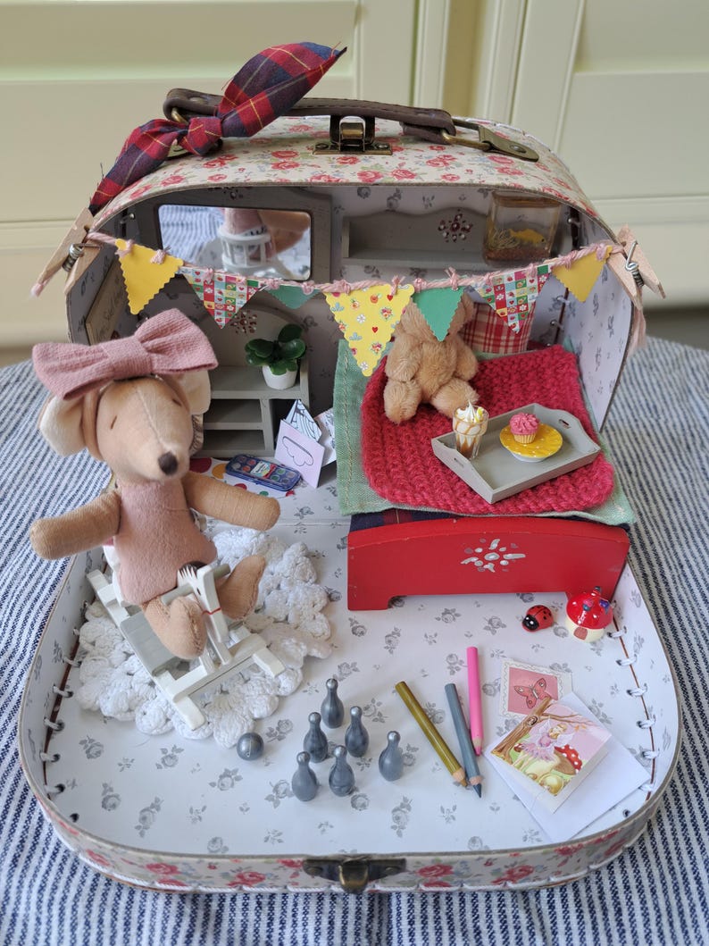 Op de afbeelding: Een miniatuur poppenhuis koffer met een bloemenpatroon, open om een gezellig interieur te onthullen. Een muispop met een roze strik zit in een miniatuurstoel. Het interieur omvat een bed, een teddybeer en miniatuuraccessoires zoals een dienblad met een cupcake.