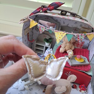 Op de afbeelding: Een miniatuur poppenhuis koffer met een bloemenpatroon, versierd met een kleurrijke banner en een kleine spiegel. Binnenin een miniatuur slaapkamer sc&egrave;ne met een bed, teddybeer en een dienblad met een cupcake. Een kleine, cr&egrave;mekleurige strik wordt voor de koffer gehouden.