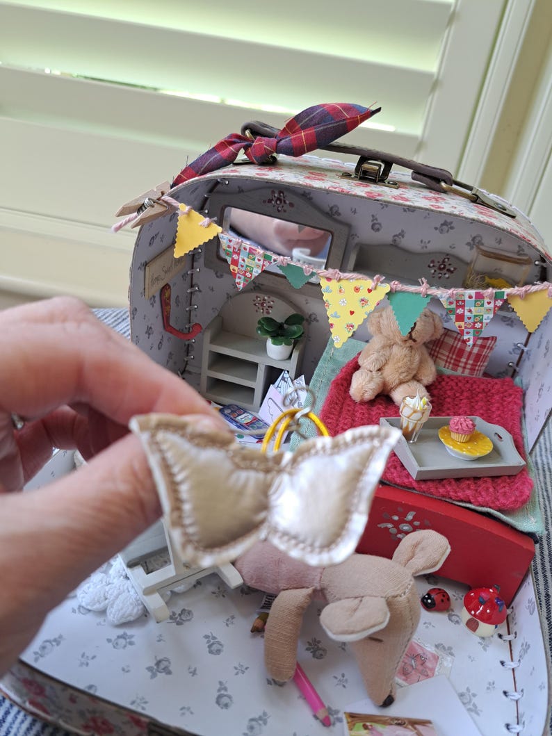 Op de afbeelding: Een miniatuur poppenhuis koffer met een bloemenpatroon, versierd met een kleurrijke banner en een kleine spiegel. Binnenin een miniatuur slaapkamer sc&egrave;ne met een bed, teddybeer en een dienblad met een cupcake. Een kleine, cr&egrave;mekleurige strik wordt voor de koffer gehouden.