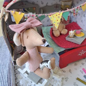 Op de afbeelding: Een gedetailleerde miniatuur poppenhuis sc&egrave;ne in een vintage koffer. Een pluchen hond met een roze jurk en strik zit op een schommelpaard. De sc&egrave;ne bevat een bed, een teddybeer en een dienblad met miniatuur desserts. Kleurrijke vlaggetjes versieren het interieur.
