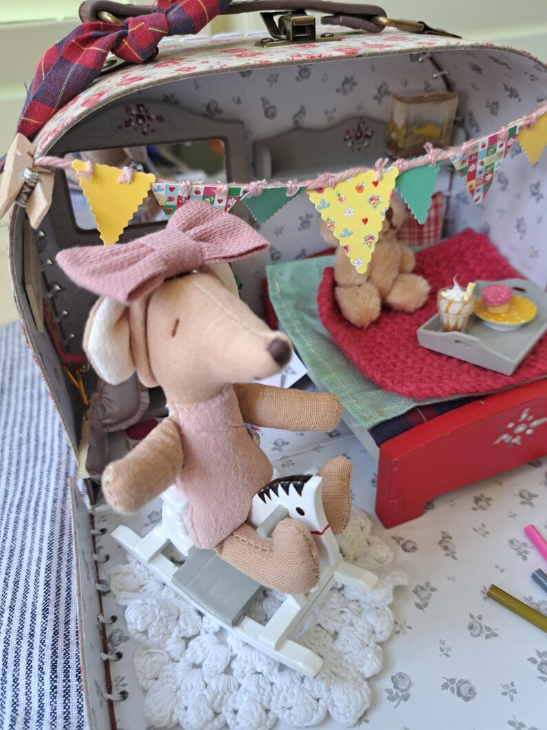 Op de afbeelding: Een gedetailleerde miniatuur poppenhuis sc&egrave;ne in een vintage koffer. Een pluchen hond met een roze jurk en strik zit op een schommelpaard. De sc&egrave;ne bevat een bed, een teddybeer en een dienblad met miniatuur desserts. Kleurrijke vlaggetjes versieren het interieur.