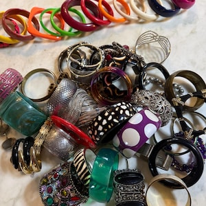 May include: A large collection of assorted bangles and bracelets in various colors, patterns, and materials. The pile includes solid-colored plastic, metallic, beaded, and patterned bangles, featuring designs like polka dots, animal prints, and rhinestones.