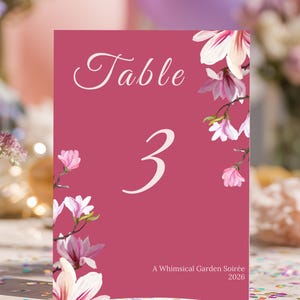 Peut inclure: Une carte de numéro de table magenta avec le mot "Table" en écriture blanche et le chiffre "3". La carte est ornée de fleurs de magnolia à l'aquarelle et du texte "A Whimsical Garden Soirée 2026".