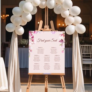 以下が含まれることがあります： 木製のイーゼルに飾られた結婚式の座席表。テキスト「Find your Seat」と花の装飾が施されています。白い風船のアーチが看板の上にあります。看板は薄いピンク色で、ゲストの名前が記載されています。クリーム色の布が両側に垂れ下がっています。