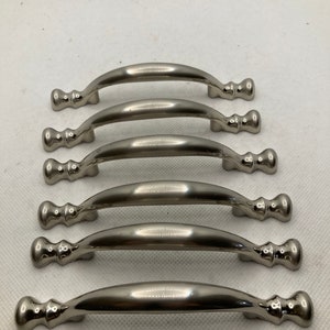 May include: Six brushed nickel cabinet pulls. Each pull has a curved handle and decorative end pieces. The hardware is designed for easy installation and adds a modern touch to any cabinet or drawer.