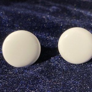 May include: Two white, round stud earrings. The earrings are smooth and glossy, with a simple, minimalist design. They are set against a dark blue, textured background.