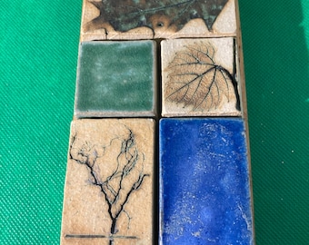 May include: A rectangular ceramic wall hanging with a gold ring for hanging. The piece is divided into six squares, each with a different design: a leaf, a tree, and solid green and blue tiles. The leaves are brown and the tree is black.