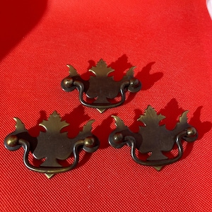 May include: Three antique brass drawer pulls with a leaf design. Each pull has a curved handle and is mounted on a decorative backplate. The pulls are arranged on a red surface.