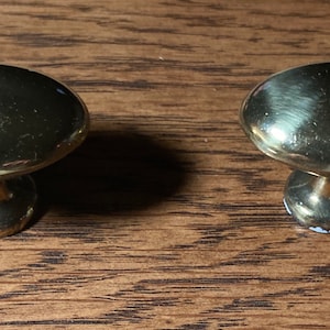 May include: Two shiny, gold-colored cabinet knobs with a rounded top and a short, cylindrical base. The knobs are set against a wooden surface, showcasing their reflective quality and design. These knobs are suitable for furniture restoration or new builds.