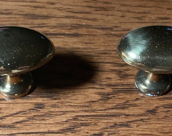 May include: Two shiny, gold-colored cabinet knobs with a rounded top and a short, cylindrical base. The knobs are set against a wooden surface, showcasing their reflective quality and design. These knobs are suitable for furniture restoration or new builds.