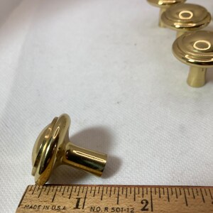 以下が含まれることがあります： 丸みを帯びた溝のデザインの、金色のキャビネットノブのコレクション。ノブは木製の定規で示されており、ノブの長さは約2.5cmです。ノブは白い表面にあります。