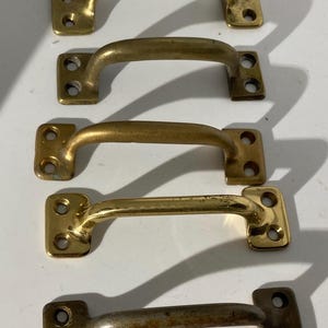 May include: Four brass-toned cabinet pulls with a curved handle design. Each pull has a rectangular base with two screw holes. The pulls vary slightly in color, with some showing signs of age and wear.