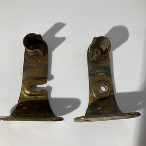May include: Two vintage brass-toned metal curtain rod brackets. Each bracket has a curved top, a vertical support, and a flat base with screw holes. The metal shows signs of age and wear, with some discoloration.