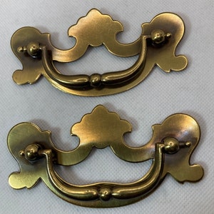 May include: Two antique brass drawer pulls with a decorative, ornate design. Each pull features a curved handle and decorative backplates with a floral-like shape. The hardware is a warm, aged brass color, suggesting vintage or retro style.