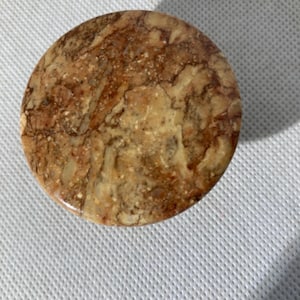 May include: A round, stone coaster with a marbled pattern in shades of brown, tan, and cream. The coaster has a smooth, polished surface and is likely made of natural stone. This coaster is suitable for use with glasses and mugs.