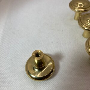 以下が含まれることがあります： 丸く平らな上面と円筒形のベースを備えた、いくつかの金色のキャビネットノブ。ノブは光沢のある金属素材でできており、引き出しやキャビネットに簡単に取り付けられるように設計されています。白い背景に表示されています。