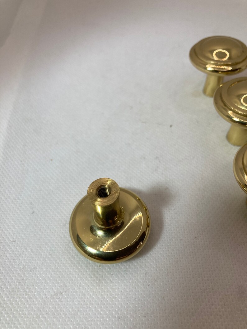 以下が含まれることがあります： 丸く平らな上面と円筒形のベースを備えた、いくつかの金色のキャビネットノブ。ノブは光沢のある金属素材でできており、引き出しやキャビネットに簡単に取り付けられるように設計されています。白い背景に表示されています。