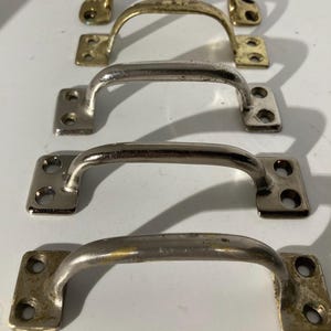 May include: Four metal cabinet handles in varying finishes: gold, silver, and tarnished brass. Each handle has a curved bar and two rectangular mounting plates with screw holes. The handles are arranged vertically against a white background.