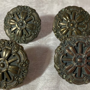 May include: Four antique bronze-colored cabinet knobs. Each knob features a detailed floral and scroll design with a central flower and radiating petal-like elements. The knobs are round and have a vintage aesthetic.