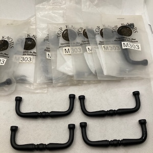 May include: A collection of black cabinet pulls and handles. Some are packaged in clear plastic with the text "TOP KNOBS" and "M303". The handles have a curved design with decorative details, suitable for furniture or cabinetry.
