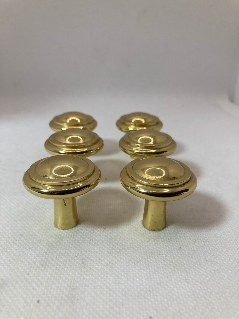 以下が含まれることがあります： キノコ型のデザインの、金色のキャビネットノブが6つ。ノブは、丸みを帯びた上部と円形の溝、円筒形のステムを備えています。ノブは3つずつ2列に配置されています。