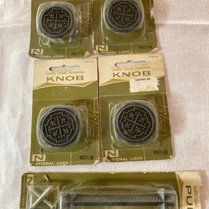 May include: A collection of vintage cabinet hardware, including four dark gray knobs with a rope-like border and a decorative center, and one matching pull handle. The packaging reads "Quality Cabinet Accessories" and "NATIONAL LOCK".