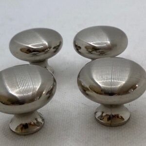 May include: Four brushed nickel cabinet knobs with a rounded, oval shape. The knobs have a smooth, reflective surface and are mounted on a short, cylindrical base. The hardware is designed for use on drawers or cabinet doors.