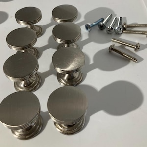 May include: A collection of brushed nickel cabinet knobs with matching screws. The knobs have a circular, flat top and a decorative base. The screws are of varying sizes and head styles. The hardware is designed for furniture or cabinetry.