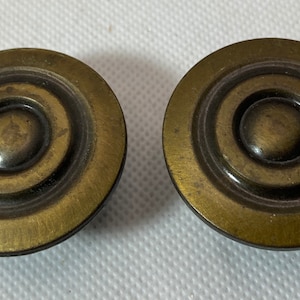 May include: Two antique brass-toned cabinet knobs. Each knob is circular with a raised, concentric design. The knobs have a vintage aesthetic, with a slightly worn appearance. These knobs are suitable for furniture restoration projects.