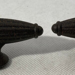 May include: Two dark brown cabinet pulls with a textured, ribbed oval design. The pulls have a decorative, rounded end and a curved base. These decorative pulls are ideal for adding a rustic touch to furniture.