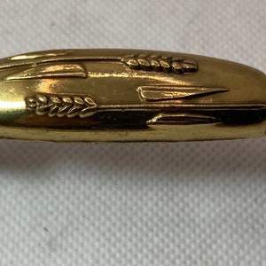 May include: A gold-colored cabinet pull with a wheat and arrow design. The central handle has a rounded shape with etched wheat and arrow details. The ends feature a clover-shaped design.