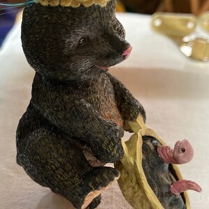May include: A whimsical figurine of a black bear wearing a decorative hat with blue feathers. The bear is holding a pod with a pink worm inside. The pod rests on a pink wheel. A charming decorative item.