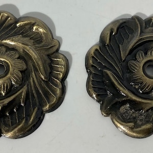 May include: Two antique bronze-colored floral decorative hardware pieces. Each piece features a central flower design surrounded by swirling leaves. The hardware has a hole in the center and is likely used for furniture or decorative purposes.