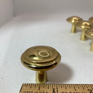 以下が含まれることがあります： 金色のキャビネットノブのセット。ノブは、装飾的なリングと円筒形のステムを備えた丸みを帯びた上部を備えています。インチの測定値を持つ木製の定規が前景にあります。ノブは白い表面にあります。