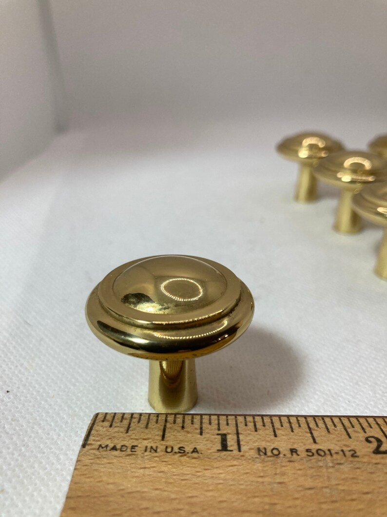 以下が含まれることがあります： 金色のキャビネットノブのセット。ノブは、装飾的なリングと円筒形のステムを備えた丸みを帯びた上部を備えています。インチの測定値を持つ木製の定規が前景にあります。ノブは白い表面にあります。