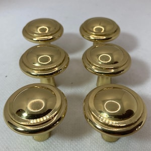 May include: Six gold-colored cabinet knobs with a rounded, tiered design. The knobs have a shiny, reflective surface and a short, cylindrical base. The knobs are arranged in two rows of three.