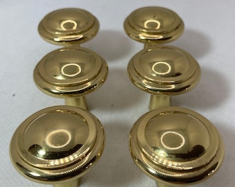 May include: Six gold-colored cabinet knobs with a rounded, tiered design. The knobs have a shiny, reflective surface and a short, cylindrical base. The knobs are arranged in two rows of three.