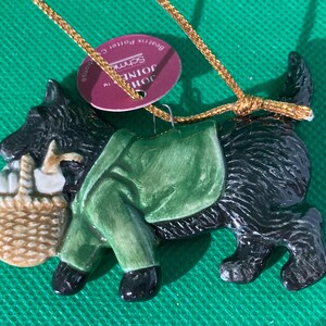 May include: A ceramic ornament in the shape of a black Scottish Terrier dog carrying a basket and wearing a green coat. The ornament has a gold string for hanging and a tag that reads "John Schmeisser".