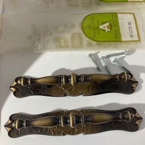 May include: Two antique bronze cabinet pulls with decorative detailing. The pulls have a curved handle and ornate end pieces. Includes mounting screws and packaging from The Carriage House Collection.