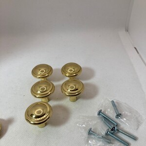 以下が含まれることがあります： 丸みを帯びた段付きデザインの、金色のキャビネットノブ6個セット。ノブは3つずつ2列に配置され、近くには小さなネジのパックが見えます。ノブは光沢のある反射面を持っています。